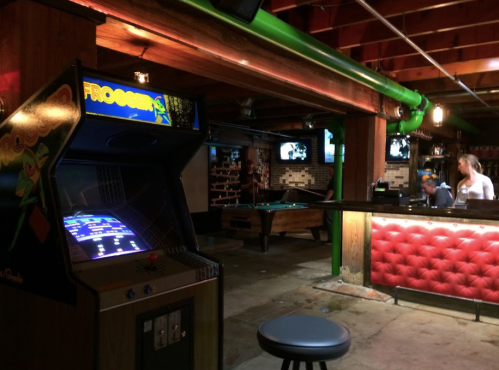 A retro arcade with a Frogger machine, a pool table, and a bar area in a dimly lit basement setting.