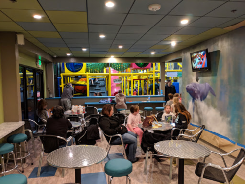A colorful indoor play area with children playing in the background, while families sit at tables in the foreground.