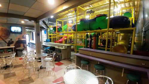 Indoor play area with colorful slides and tunnels, visible from a seating area with metal tables and chairs.