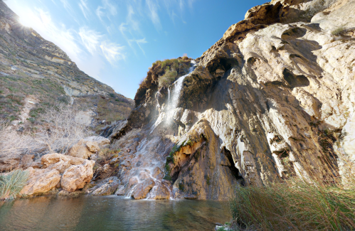 A serene waterfall cascading down rocky cliffs into a calm pool, surrounded by lush vegetation and blue sky.