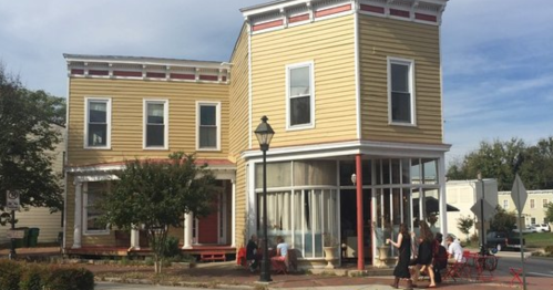A two-story yellow building with large windows, featuring a corner entrance and people sitting outside on a sunny day.