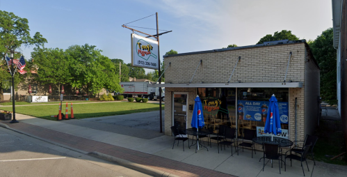 A small restaurant with outdoor seating, blue umbrellas, and a "Now Hiring" sign, surrounded by greenery.