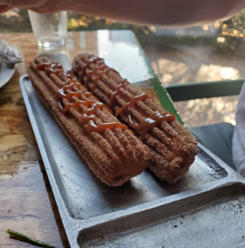 Two churros drizzled with caramel sauce on a metal tray, with a glass in the background.