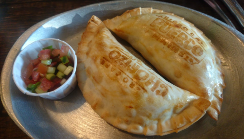 Two golden-brown empanadas on a metal plate, accompanied by a small cup of fresh salsa.