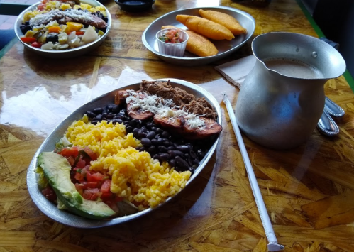 A platter of rice, black beans, avocado, tomatoes, and meat, with a pitcher of drink and a side of pastries.