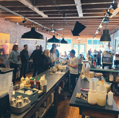 A busy café interior with customers at the counter and people seated, featuring exposed brick and wooden beams.