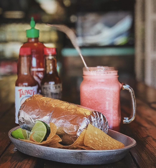 A foil-wrapped burrito on a plate with lime wedges and chips, alongside a smoothie and hot sauce bottles in the background.