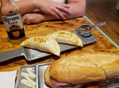 A sandwich and two empanadas on a metal tray, with a Corona beer and a small bowl of salsa in the background.