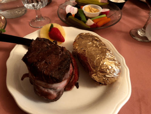 A plate with a bacon-wrapped steak, a foil-wrapped potato, and a side of fruit and vegetables.