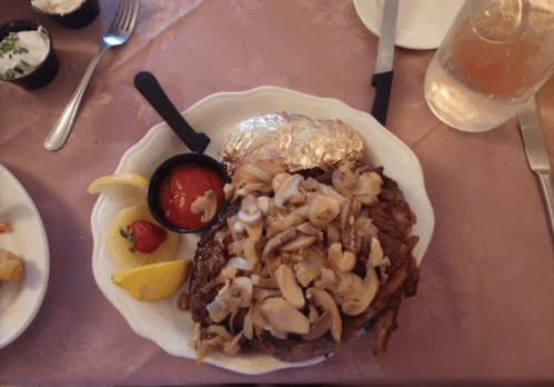 A plate featuring a large steak topped with mushrooms, served with a baked potato, lemon wedges, and a side of salsa.