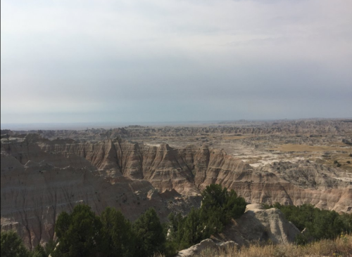 A vast, rugged landscape of layered rock formations under a cloudy sky, with sparse vegetation in the foreground.