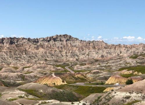 A winding road cuts through rugged, colorful rock formations under a clear blue sky in a vast landscape.