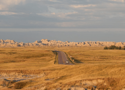 A winding road stretches through golden grasslands, leading towards rugged, layered rock formations under a blue sky.