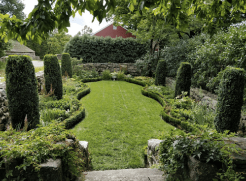 Lush garden with manicured hedges, stone walls, and a green lawn, surrounded by trees and a red barn in the background.