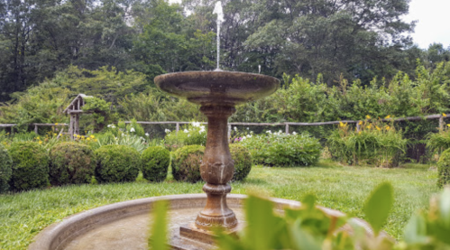A stone fountain with a gentle water spray, surrounded by lush greenery and colorful flowers in a garden setting.