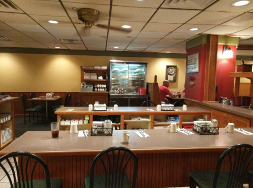 A cozy diner interior with wooden tables, chairs, and a glass display case, featuring a few patrons and condiments on tables.