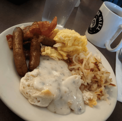 A plate of breakfast featuring scrambled eggs, bacon, sausage, hash browns, and biscuits with gravy, alongside a coffee mug.