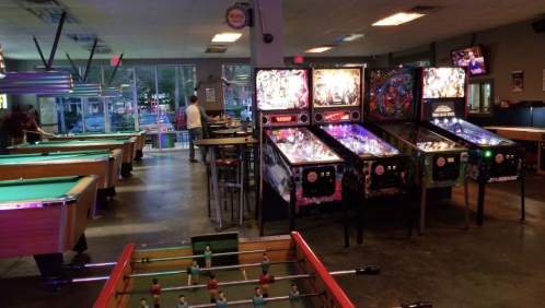 A game room featuring pool tables, pinball machines, and a foosball table, with dim lighting and a lively atmosphere.