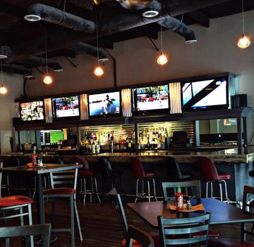 A modern bar interior with multiple TVs, a well-stocked bar, and red chairs at tables under warm pendant lighting.