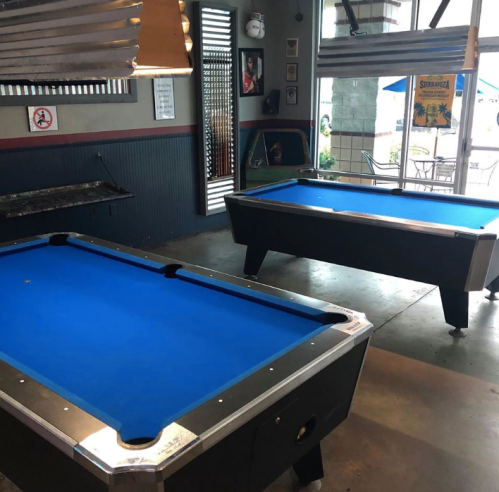 Two blue pool tables in a casual indoor setting with a bar and outdoor seating visible through a window.