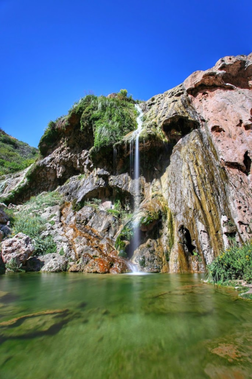 A serene waterfall cascading over rocky cliffs into a clear green pool, surrounded by lush vegetation and a blue sky.