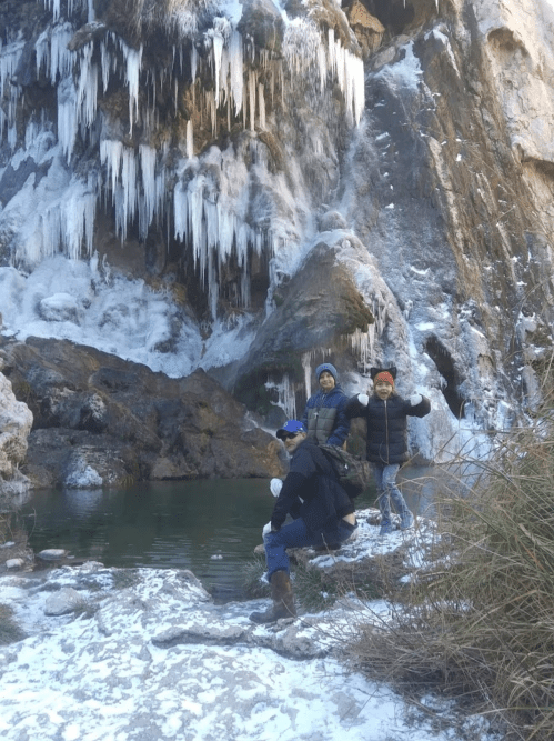 Three people pose near a frozen waterfall, surrounded by ice and snow, with a small pool of water in the foreground.