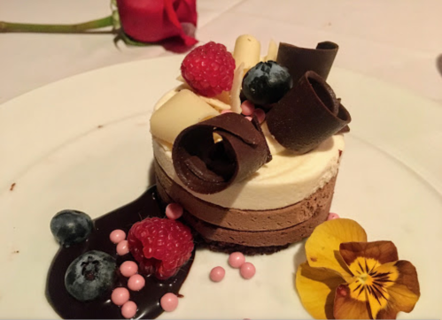 A decadent chocolate mousse cake topped with raspberries, blueberries, chocolate curls, and edible flowers on a plate.