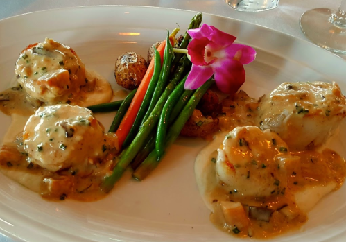 A plate of scallops in creamy sauce, served with green beans, roasted potatoes, and a purple orchid garnish.