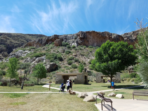 A scenic park with green grass, trees, and rocky cliffs in the background; people walking and enjoying the outdoors.