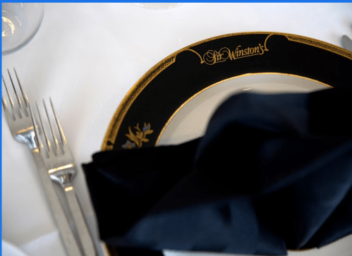A black plate with "Winston's" in gold, accompanied by a folded black napkin and silver cutlery on a white tablecloth.