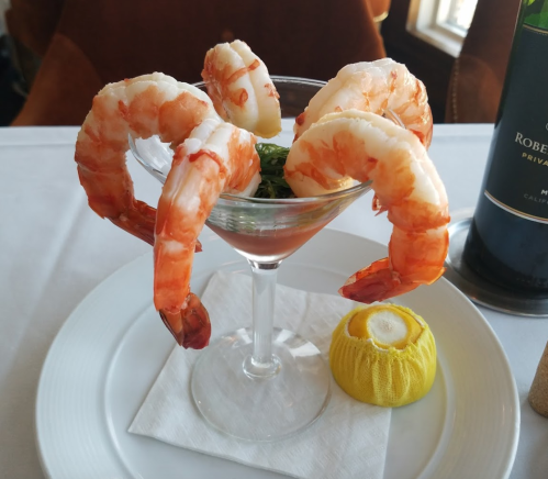 A shrimp cocktail in a glass, featuring large shrimp arranged around a bed of greens, with a lemon wedge on the side.