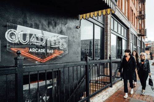 Two people walk past a neon sign for "Quarters Arcade Bar" on a city street.