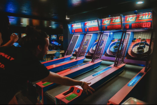 A person playing an arcade game, aiming at a colorful ice ball machine with glowing screens in a dimly lit arcade.