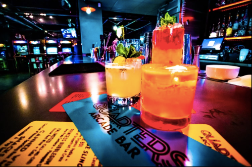 Colorful cocktails on a bar with arcade games in the background, featuring vibrant lighting and a menu.