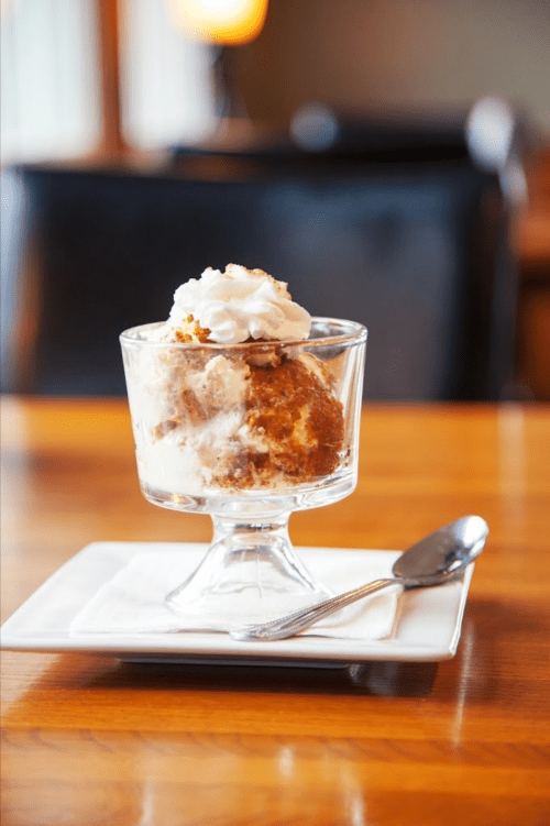 A glass dessert dish filled with ice cream and cake, topped with whipped cream, on a white plate with a spoon.