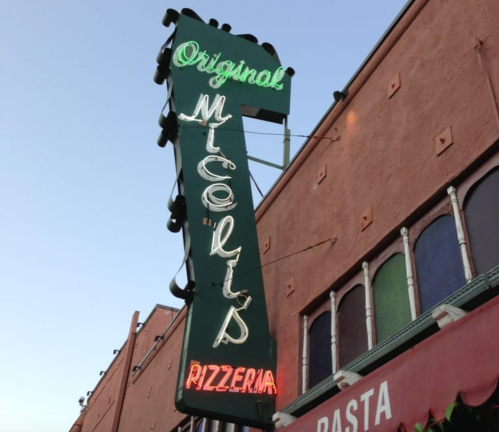 Neon sign for "Original Miceli's Pizzeria" against a colorful building backdrop.