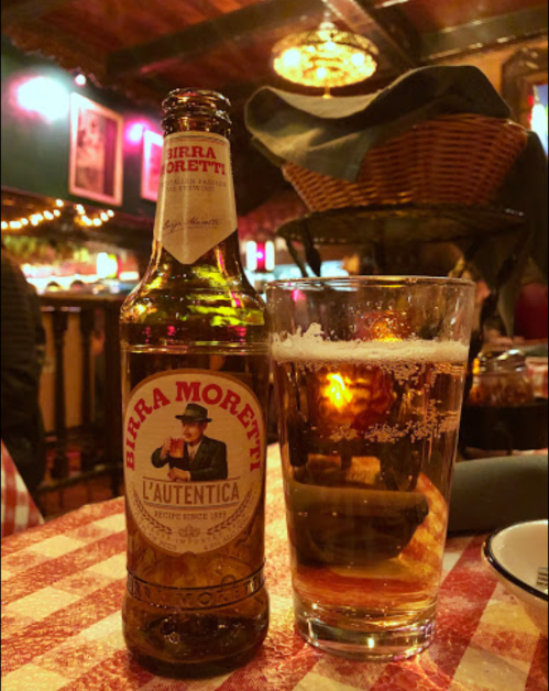 A bottle of Birra Moretti next to a glass of beer on a checkered tablecloth in a cozy restaurant setting.