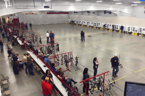 A spacious indoor archery range with rows of targets, archers, and spectators seated in a large, well-lit area.