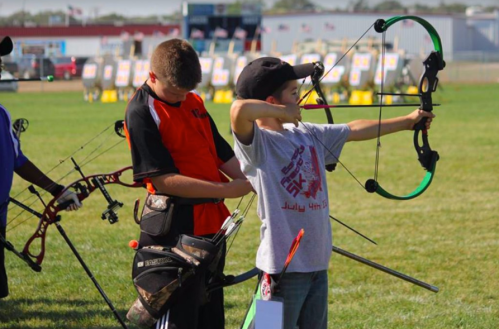Two young archers focus on their targets during an outdoor archery event, with colorful bows and arrows in hand.