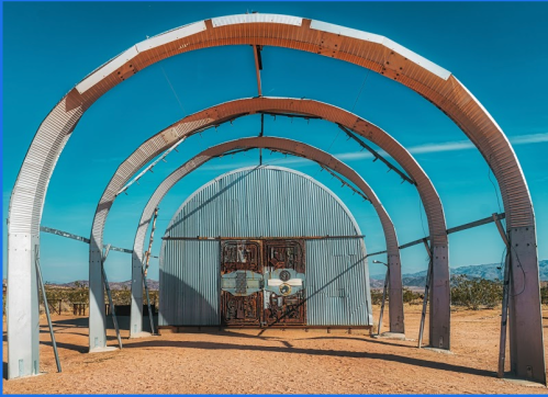 A large, metallic structure with arched frames and a central door, set against a clear blue sky in a desert landscape.