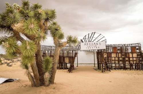 Abandoned outdoor theater structure with a sign reading "Adrian 5 Little Theatre," surrounded by desert landscape and Joshua trees.