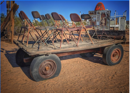 A wooden cart with stacked metal chairs on a sandy surface, set against a clear blue sky.