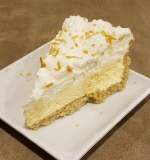 A slice of creamy yellow pie topped with whipped cream and toasted coconut flakes on a square white plate.