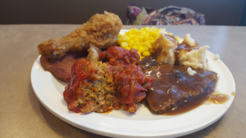 A plate of food featuring fried chicken, corn, mashed potatoes with gravy, meatloaf, and a side of tomato-based stuffing.