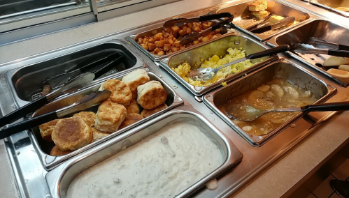 A buffet spread featuring biscuits, scrambled eggs, potatoes, and gravy in metal trays with serving utensils.