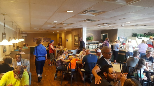 A busy restaurant interior with diners at tables, a buffet area, and staff serving customers.