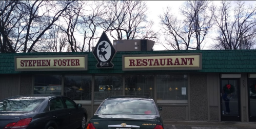 Exterior of Stephen Foster Restaurant, featuring a green roof and a sign with a silhouette logo. Cars parked in front.