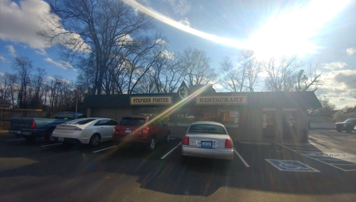A restaurant building with a sign reading "Stephen Foster" and parked cars in front, under a bright sky.