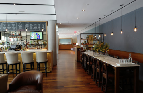 Modern restaurant interior featuring a bar, wooden floors, and a long dining table with stylish lighting.