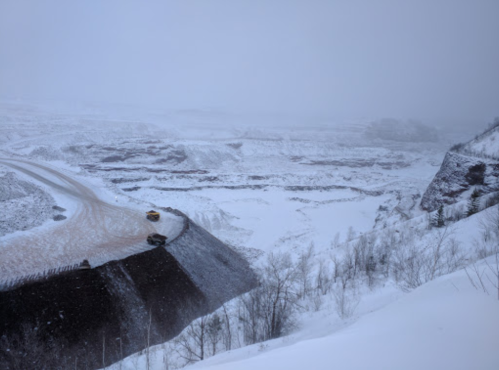 A snowy landscape with a winding road and vehicles near a steep, icy cliff in a foggy atmosphere.